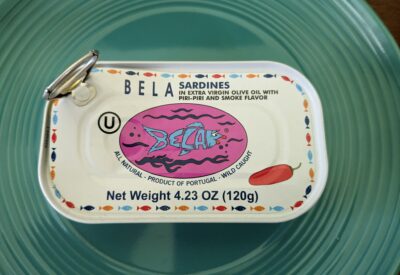 An unopened tin of Bela sardines with piri-piri and smoke flavor sitting on a blue-green plate.